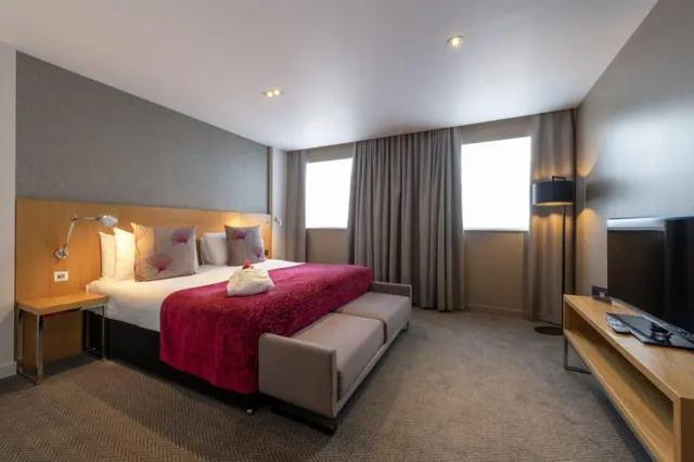 A hotel bedroom with a king bed under a red throw, an upholstered bench at the foot of the bed, a wall-mounted reading lamp and a television on a wooden stand.