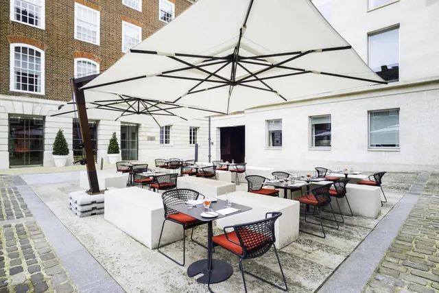 An outdoor courtyard dining area with large cream parasols, black mesh chairs with red cushions and tables laid for service, beside brick and stone buildings.