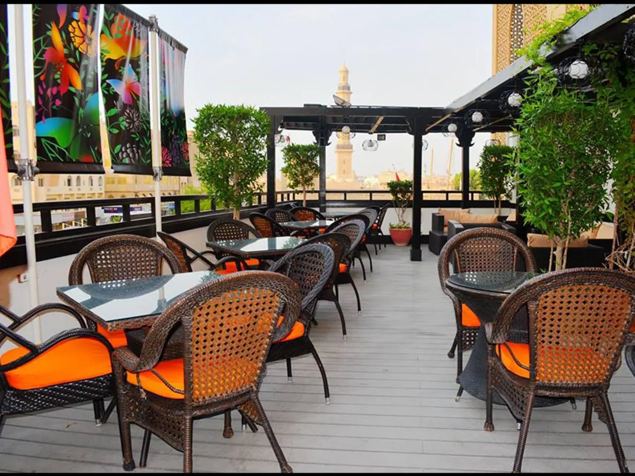 A rooftop terrace with wicker chairs with orange cushions and glass-topped tables, with a minaret tower visible in the background.