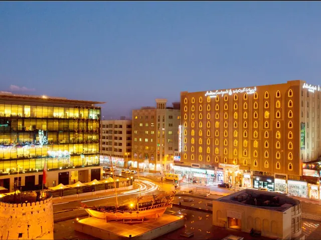 Dusk photo of an illuminated hotel building with arched windows, a street with traffic light trails, a dhow sculpture on a plinth and a fortress-style wall at left.