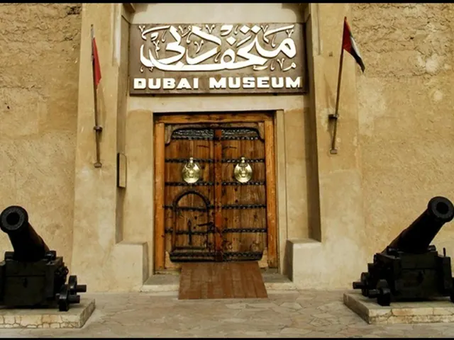 The entrance of the Dubai Museum shows a large wooden double door under a lit sign, flanked by two cannons and two flags on a sandy-coloured stone facade.