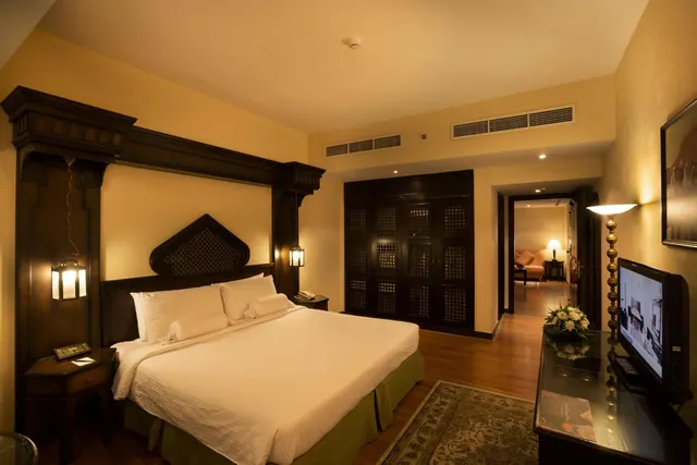 A bedroom with a king bed under a carved dark wood headboard, a latticed wooden wardrobe, a wall-mounted television and a doorway through to another room.