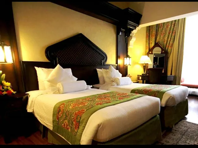 Two single beds with green and orange patterned runners sit in a warmly lit hotel room beside a desk, mirror and a window with curtains.