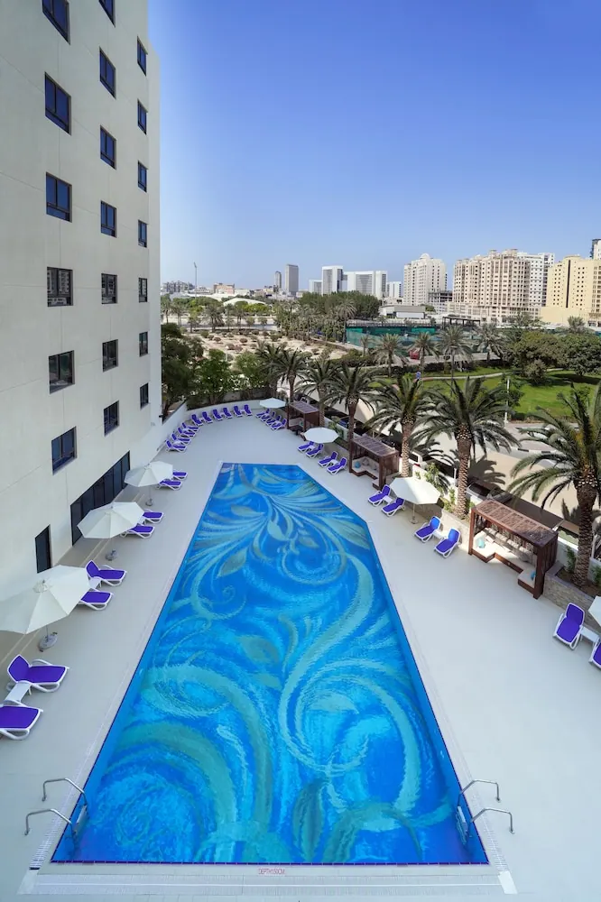 An elevated view of a long rectangular pool with a blue mosaic floor, surrounded by purple sun loungers, white umbrellas and palm trees, with a city skyline behind.