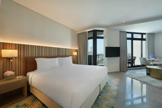 A king bed with white linen sits in a hotel room beside a seating area, a television, and glass doors opening onto a balcony with a view of distant city buildings.