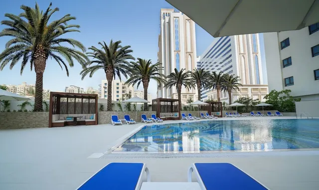 An outdoor pool with blue sun loungers, shaded cabanas and palm trees, with high-rise towers visible behind.