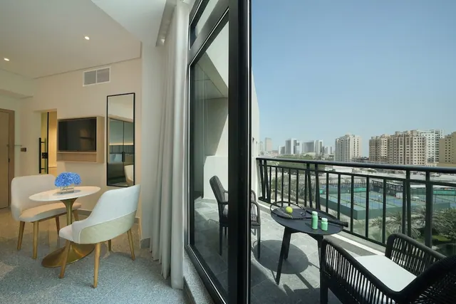 A hotel room with a round table and upholstered chairs and a wall-mounted television, with a sliding glass door opening onto a balcony with black woven chairs and a view of city towers and tennis courts.