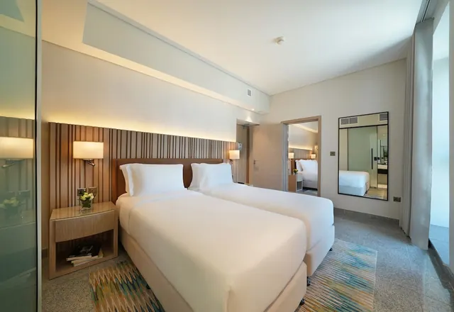A bedroom with two single beds with white bedding, a wood slat headboard wall, a bedside table with flowers and books, and a wall mirror beside an open doorway.