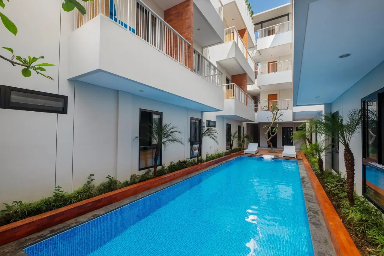 A long blue swimming pool runs through a courtyard lined with palms and planting, with white multi-storey buildings and balconies on either side and two sun loungers at the far end.