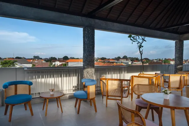 A covered rooftop terrace with rattan chairs, round wooden tables and blue armchairs, overlooking rooftops and trees under a partly cloudy sky.
