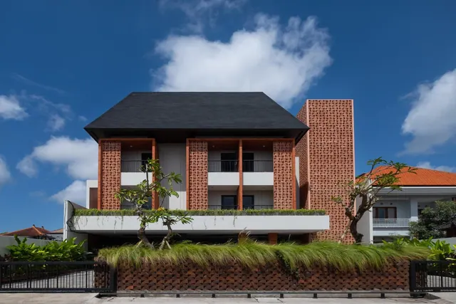 Front view of a two-storey building with a dark pitched roof, perforated brick walls, upper-floor balconies and a planted fence under a blue sky.