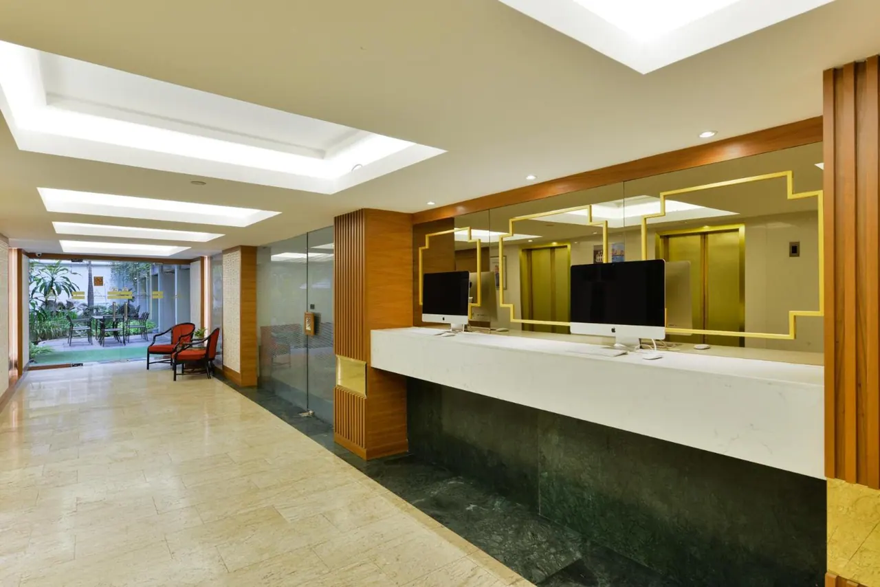 A hotel reception counter with two monitors sits under a gold-framed mirror that reflects lift doors, with a garden courtyard and red chairs visible through the opening on the left.