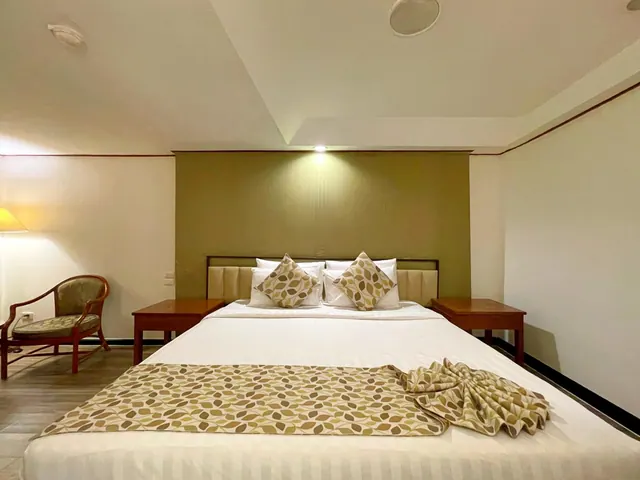 A double bed with white linens, leaf-patterned runner and matching cushions sits between two wooden nightstands, with an armchair and a lit floor lamp at the left.