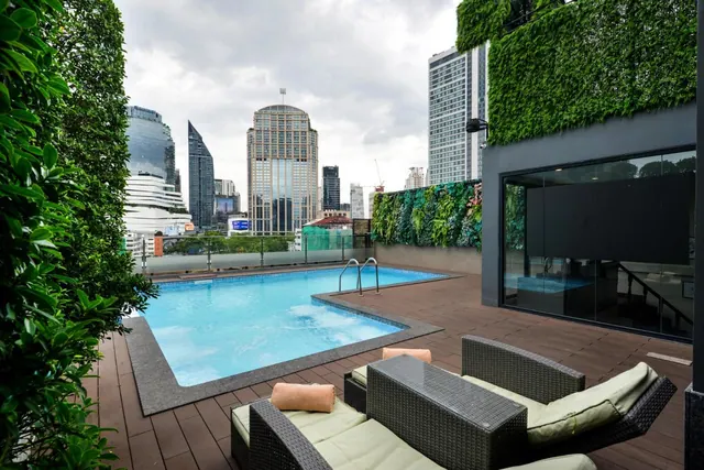 A rooftop swimming pool with wicker loungers on timber decking, with high-rise towers and green living walls beyond.
