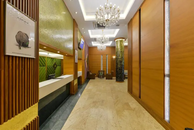 A hotel lobby with a reception desk against a green padded wall, several chandeliers hanging from the ceiling, and a seating area with armchairs at the far end.