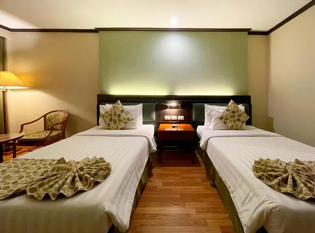 Two twin beds with patterned folded runners and cushions face a green headboard wall, with a wooden nightstand between them and a patterned armchair and floor lamp at the left.