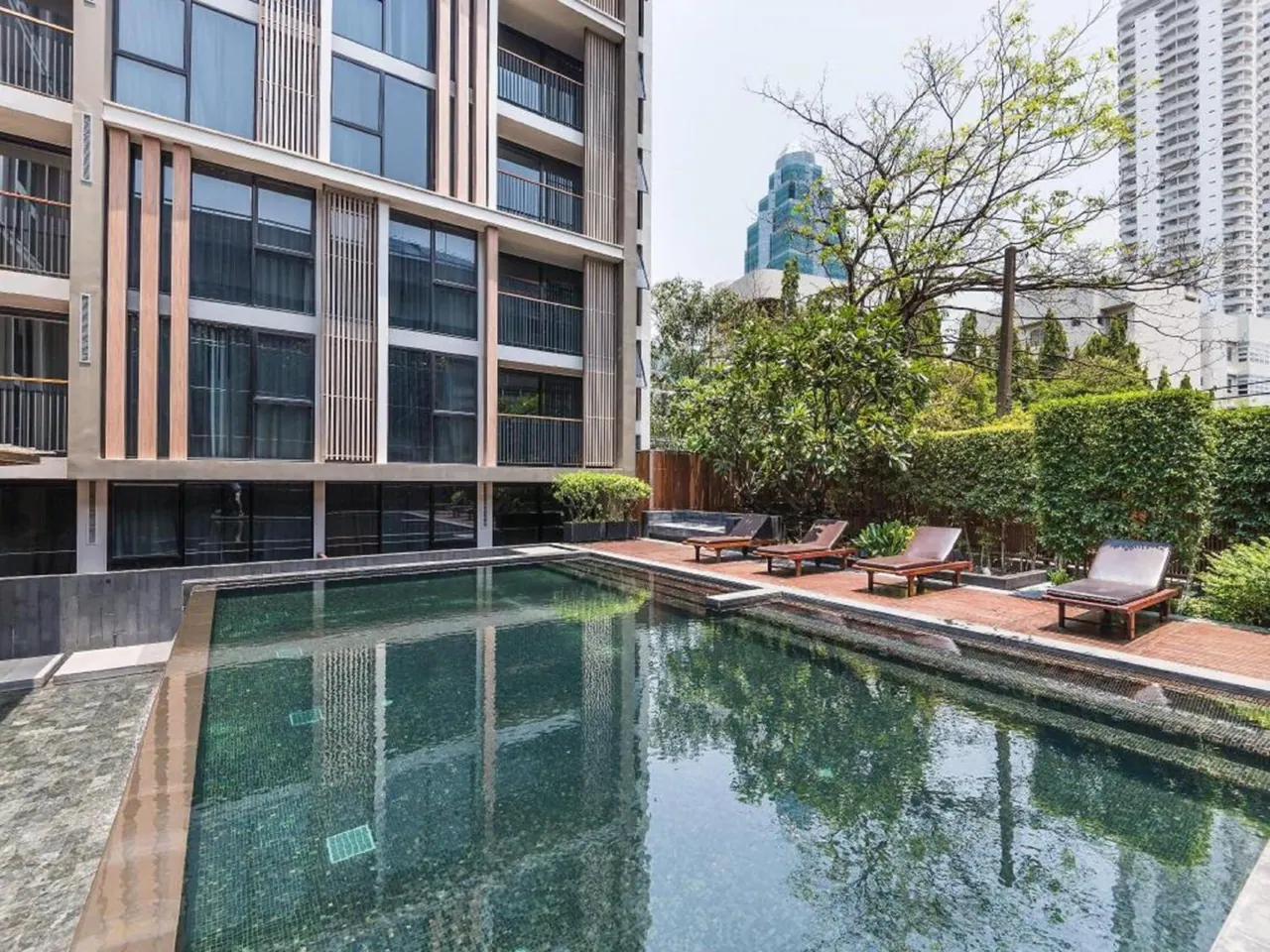 A rectangular swimming pool sits beside a tiled deck with several wooden sun loungers, next to a multi-storey building with trees and hedges behind.