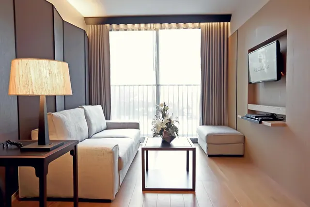 A living area with a light grey sofa, an ottoman, a wall-mounted television, a small coffee table and a table lamp, with a sliding glass door to a balcony behind.