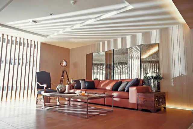 A lounge seating area with a brown leather sofa, a low metal coffee table, an upholstered armchair and mirrored panels in front of a wood-slatted wall under a striped ceiling.