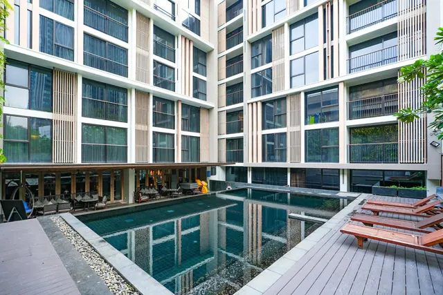 A rectangular courtyard pool sits between multi-storey building facades, with wooden sun loungers on the deck and a covered seating area under the building at left.