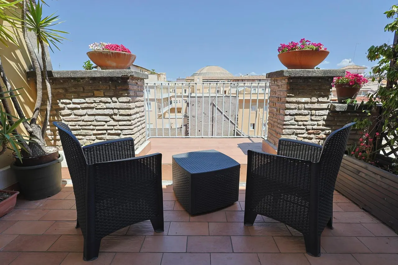 A rooftop terrace with two black wicker armchairs, a matching cube table, flower planters on stone pillars, and a view over neighbouring rooftops and a dome.