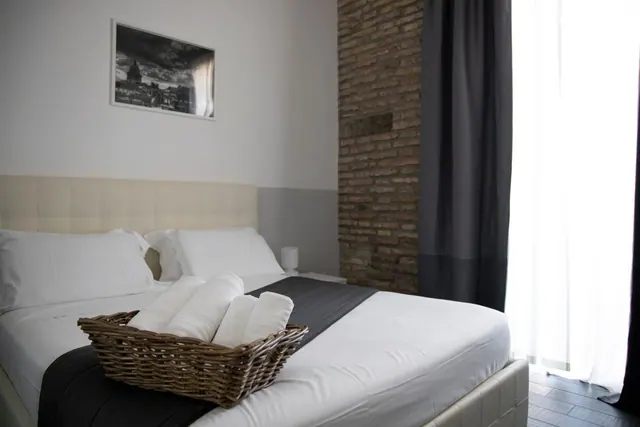 A made double bed with white linens, a dark runner and a wicker basket of folded towels, beside an exposed brick wall and a framed black and white print.