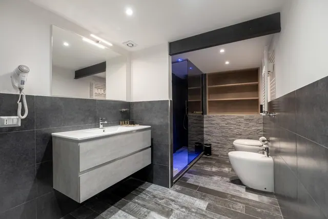 A bathroom with a wall-mounted vanity and large mirror, a hair dryer on the wall, a glass-walled shower lit in blue, and a bidet beside a toilet.