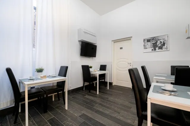 A breakfast room with small white tables, black leather chairs, a wall-mounted air conditioner and a wall-mounted television, with a door and framed black-and-white photo on the far wall.
