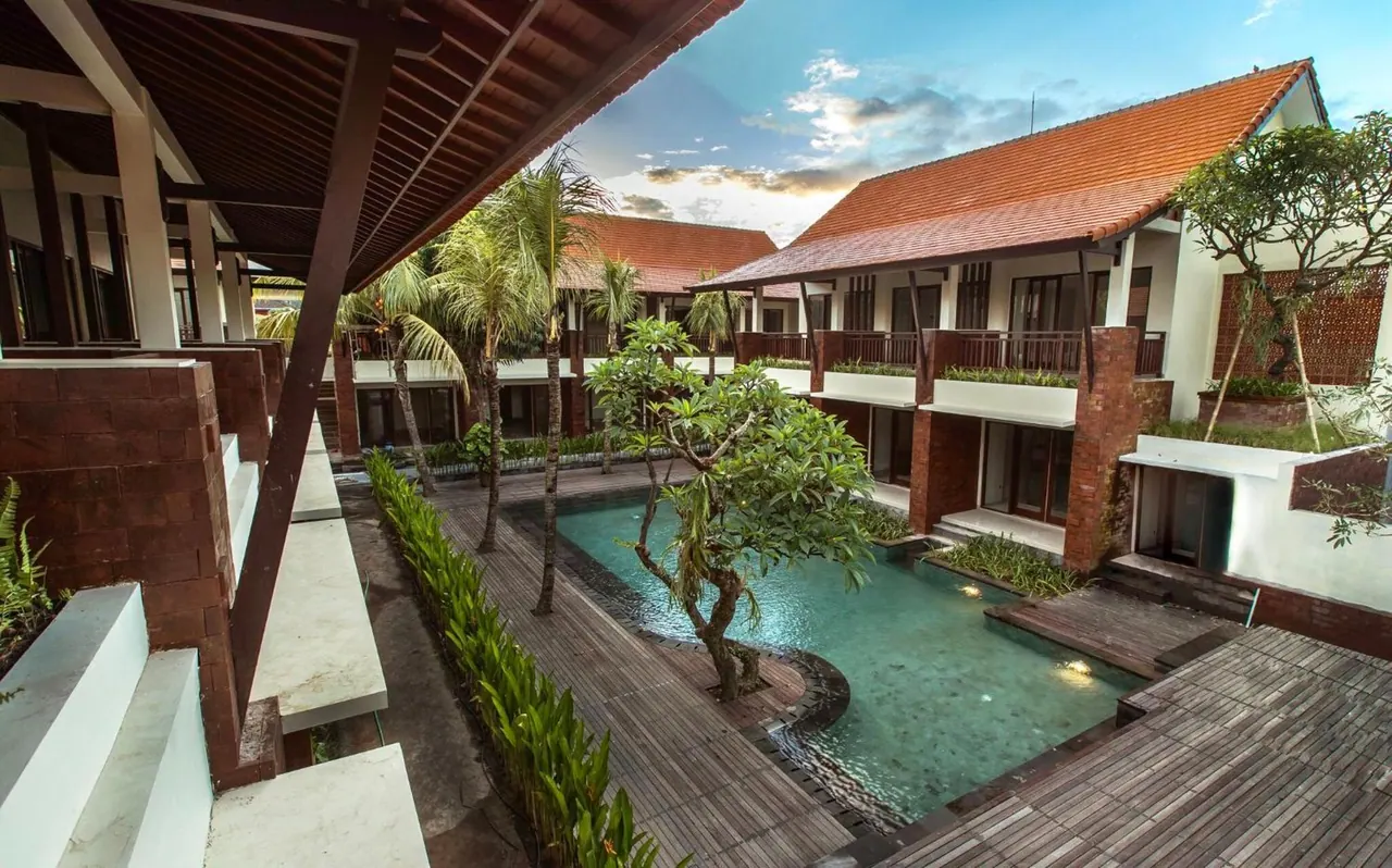 A courtyard pool with timber decking and frangipani trees, flanked by two-storey buildings with terracotta roofs and balconies under a dusk sky.