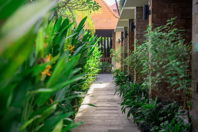 A paved pathway runs between tropical plants with orange flowers on the left and brick room buildings with wall lights on the right.