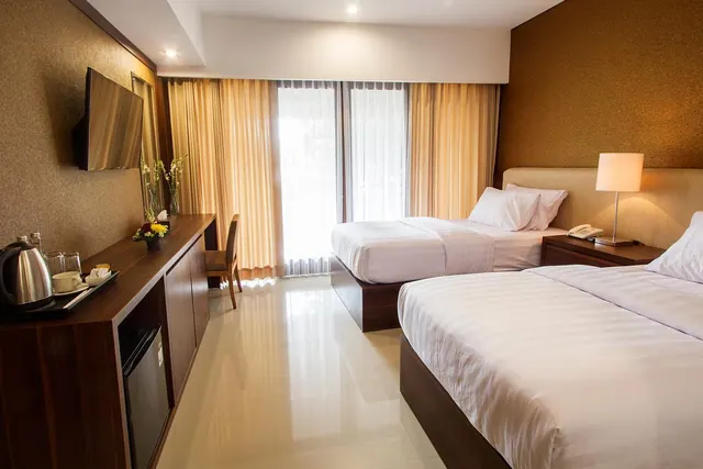 A hotel bedroom with two beds, a wall-mounted television, a tea tray with a kettle, a desk with a chair, and a minibar beneath the console, with sheer curtains over a bright window.