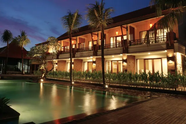A pool at dusk in the foreground, with a lit two-storey hotel building with balconies and palm trees behind it.