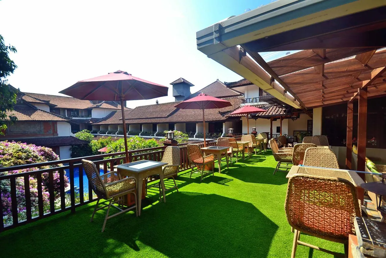 A covered terrace with green artificial turf, wicker chairs and red parasols overlooks a pool and traditional-style buildings.