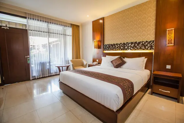 A hotel bedroom with a king bed, an armchair and small round table beside a sheer-curtained glass door, and wood-panelled walls.