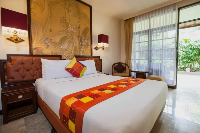 A bedroom with a double bed under a leather-tufted headboard, an armchair and small round table by a sliding glass door that opens onto greenery outside.
