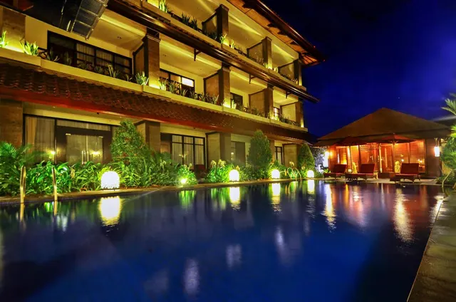 Illuminated swimming pool at night beside a multi-storey hotel building, with sun loungers and a lit pavilion to the right.