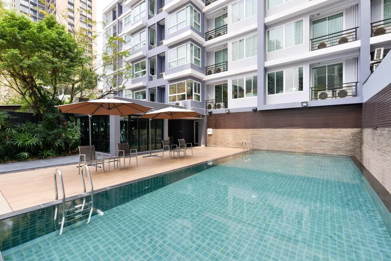 A rectangular tiled swimming pool sits beside a wooden deck with umbrellas and chairs, in front of a multi-storey building with balconies.