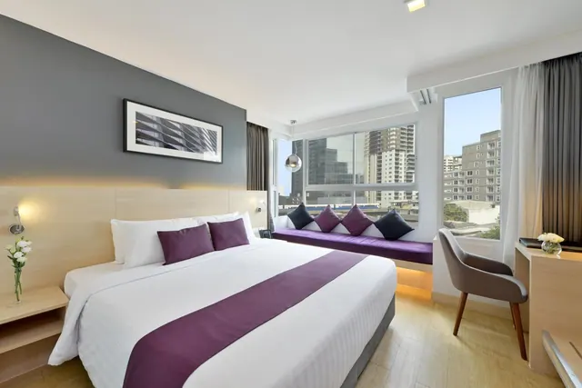 A bedroom with a king bed, a purple window seat, and a desk and chair beside a window overlooking city buildings.