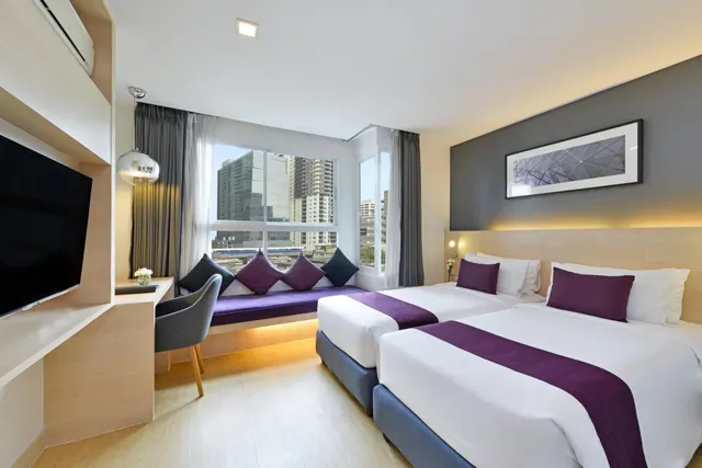 A bedroom with two twin beds, a window seat with purple cushions, a wall-mounted television, a desk, and a view of high-rise buildings through the window.