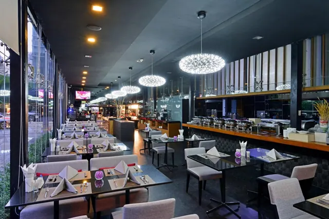 A restaurant dining room with set tables, pendant chandeliers, a buffet counter with chafing dishes along the right wall, and floor-to-ceiling windows onto a street.