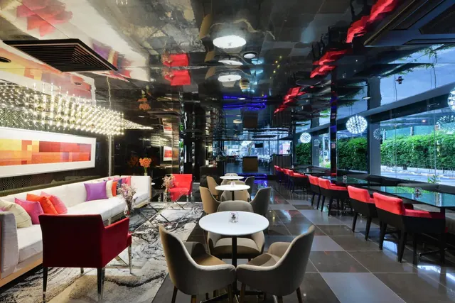 A dining room with a mirrored ceiling, red and grey chairs around round tables, a white sofa with cushions on the left, and large windows looking onto green shrubs.