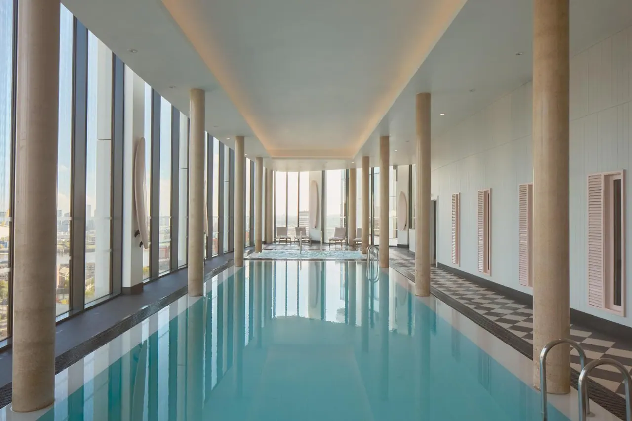 An indoor swimming pool with tall stone-look columns, floor-to-ceiling windows showing distant buildings, and a small hot tub with chairs at the far end.