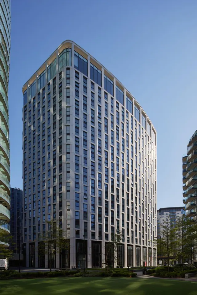 A tall modern tower with grid-patterned windows rises against a clear blue sky, with a lawn and young trees in the foreground.
