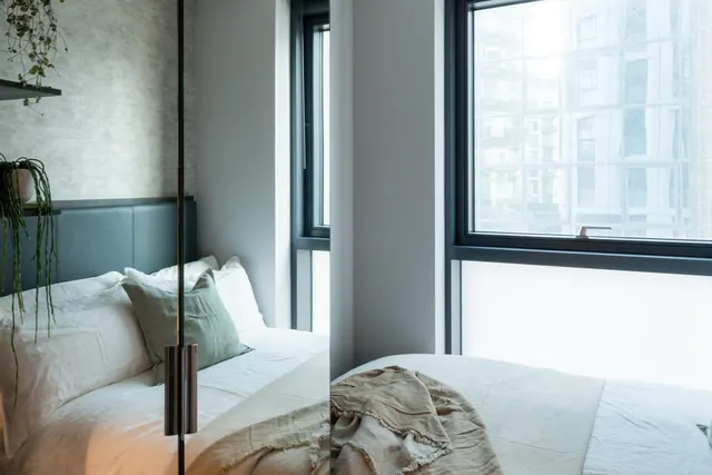 A made double bed with white linen and a sage green cushion sits beside a large window that looks onto neighbouring buildings, with a glass partition in the foreground.