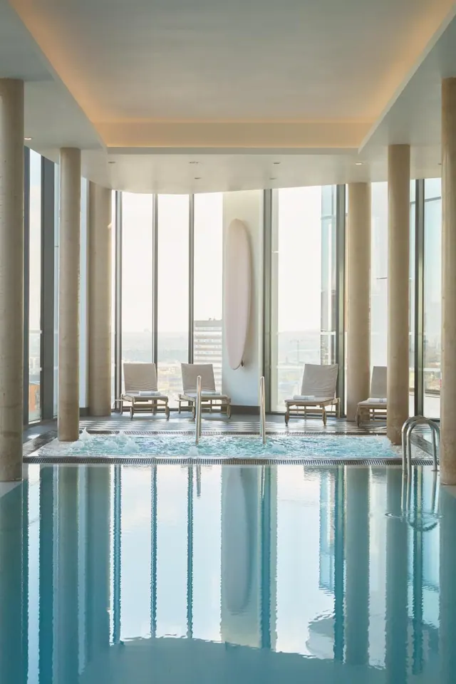 An indoor pool and a bubbling hot tub sit beside floor-to-ceiling windows, with sun loungers and a hazy city skyline beyond.
