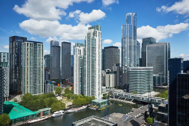 Elevated view of a skyline of tall glass and white residential towers above a dock with moored boats, a footbridge and trees under a blue sky with clouds.