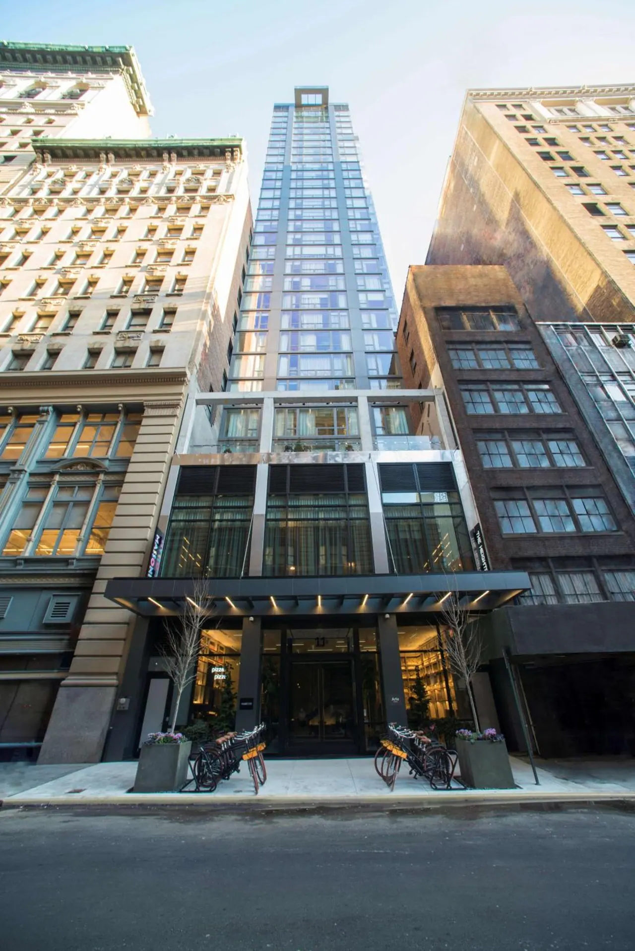 The street-level entrance of a tall glass hotel tower with a dark canopy, flanked by ornate older stone and brick buildings, with parked bicycles and planters on the sidewalk.