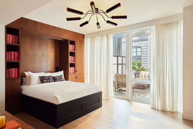 A bedroom with a made platform bed, built-in wood cabinets holding red books, and glass doors opening onto a small terrace with chairs and a potted plant.