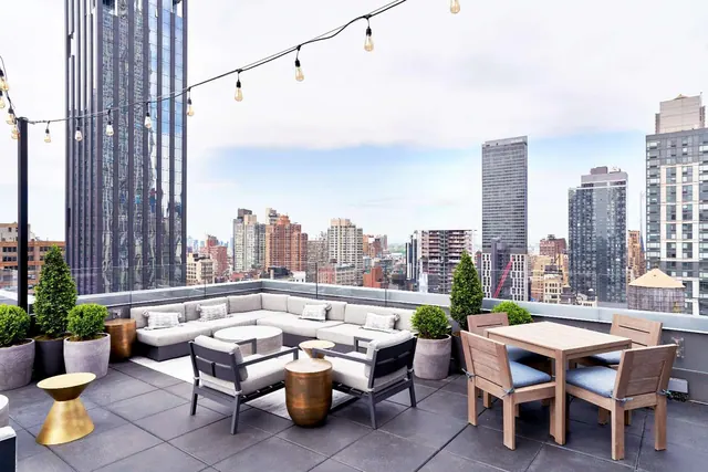 A rooftop terrace with a sectional sofa, lounge chairs, a wooden dining table and string lights, overlooking a city skyline.