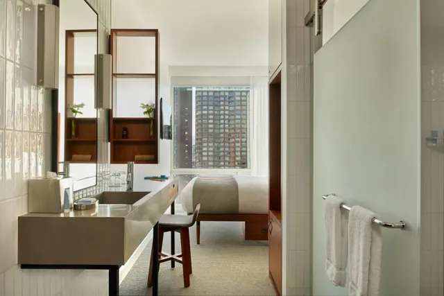 A hotel room with a double bed, a wall-mounted sink with a stool, and a window looking out over high-rise buildings.